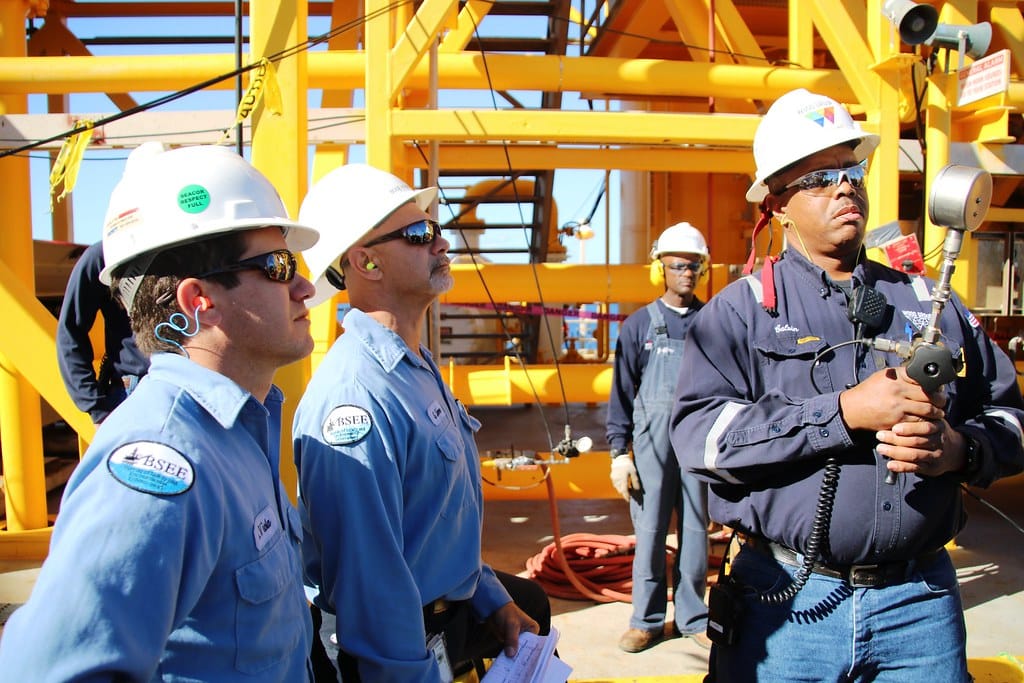 Three inspectors in hard hats on an offshore platform, one holding up a test instrument while the others watch the reading